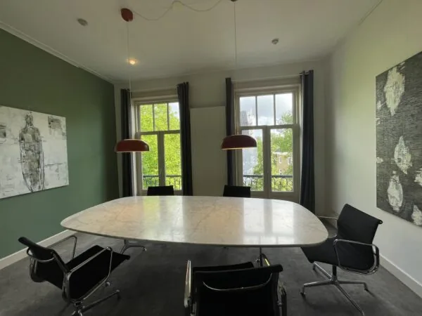 Vergaderruimte aan de Oranje Nassaulaan met ovale marmeren tafel, moderne stoelen en twee grote ramen met uitzicht op groene bomen.